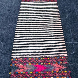 May include: A rectangular, hand-woven rug with a black and white striped pattern. The ends feature intricate, colorful geometric designs in pink, red, blue, and yellow. The rug has a fringed edge and is likely used as a decorative floor covering.