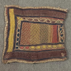 Op de afbeelding: Rechthoekig, met de hand gestikt textiel met een gefranjerde rand. Het ontwerp heeft een centraal paneel met verticale strepen en zigzagpatronen in bruin, geel, rood en groen. De rand is donkerbruin met blauwe en gele accenten.
