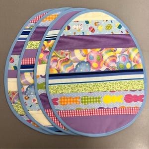 May include: Set of four purple, blue, green and white Easter egg-shaped placemats with a light blue trim. The placemats feature a variety of Easter-themed patterns, including stripes, polka dots and bunnies.