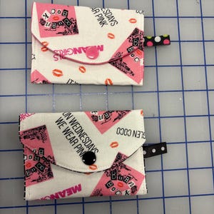 May include: Two small, rectangular fabric wallets with a white background and a pink and black design. The wallets feature text that reads "Mean Girls" and "On Wednesdays We Wear Pink." Each wallet has a button closure and a small fabric loop.