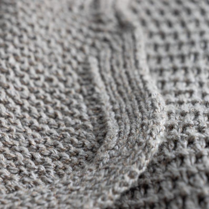May include: Close-up of a gray knitted fabric with a cable knit design. The texture is visible, showing the individual stitches and the subtle variations in the yarn's color. The fabric appears soft and warm.