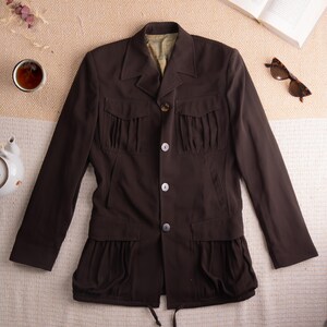 Könnte beinhalten: Dunkelbraune Jacke mit vier Fronttaschen und Knopfverschluss. Die Jacke hat ein einzigartiges Design mit plissierten Details an den Taschen und am unteren Saum. Die Jacke wird auf einer strukturierten Oberfläche präsentiert, mit einer Tasse Tee, einer Teekanne und Sonnenbrillen.