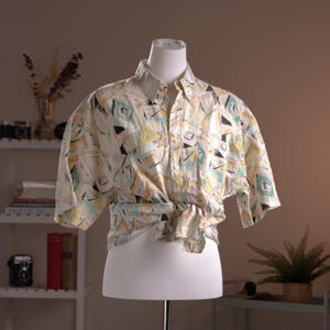 May include: A short-sleeved button-down shirt with a colorful geometric pattern in shades of cream, yellow, green, and black. The shirt is knotted at the waist and displayed on a white mannequin.