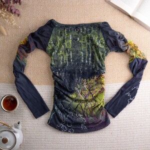 May include: A long-sleeved top with a forest-themed design in green, blue, and purple. The top has a boat neck and gathered detailing. The sleeves are navy blue with a patterned section near the wrist. A teapot and teacup are in the image.