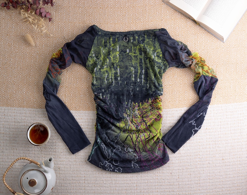 May include: A long-sleeved top with a forest-themed design in green, blue, and purple. The top has a boat neck and gathered detailing. The sleeves are navy blue with a patterned section near the wrist. A teapot and teacup are in the image.