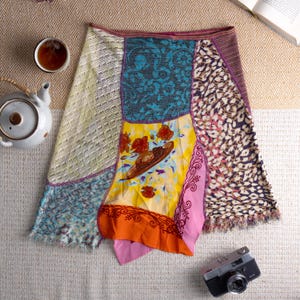Könnte beinhalten: Ein Patchwork-Rock mit verschiedenen Stoffmustern, darunter Blumen- und Tiermuster. Der Rock hat eine Mischung aus leuchtenden Farben wie Gelb, Blau und Rosa und einen Fransenbesatz. Eine Vintage-Kamera und ein Teeservice sind im Hintergrund zu sehen.