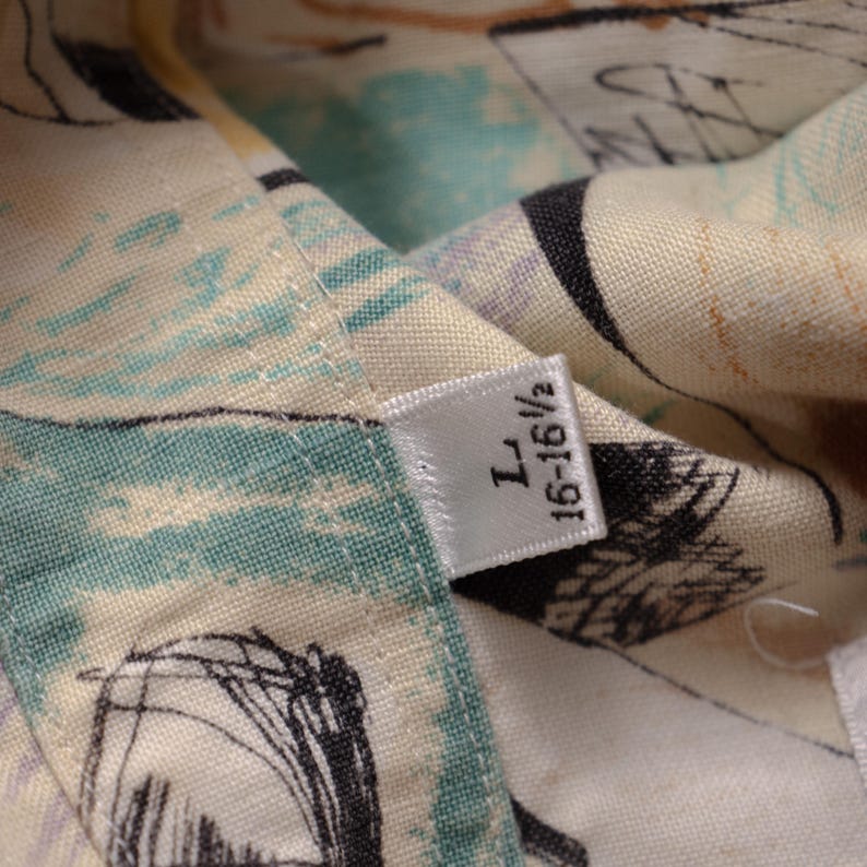 May include: Close-up of a patterned fabric with a tag. The fabric features a mix of colors, including cream, teal, and black, with abstract designs. The tag reads "L 16-16 1/2".