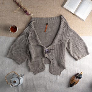 May include: A light brown, short-sleeved cardigan with a unique button closure. The sweater has a textured knit design and a wide collar. A small orange tag is attached near the collar. The cardigan is displayed on a neutral-toned surface, with a teacup and book nearby.