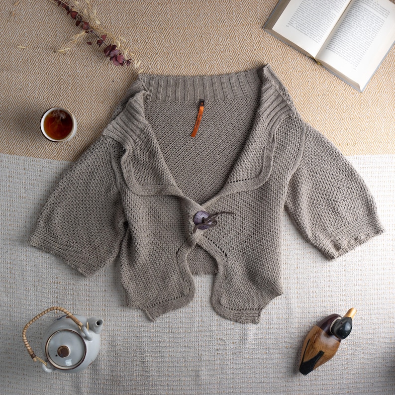 May include: A light brown, short-sleeved cardigan with a unique button closure. The sweater has a textured knit design and a wide collar. A small orange tag is attached near the collar. The cardigan is displayed on a neutral-toned surface, with a teacup and book nearby.