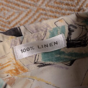 May include: Close-up of a fabric label that reads "100% LINEN." The fabric is a light cream color with a pattern of black, teal, and purple abstract shapes. The label is white with black text.