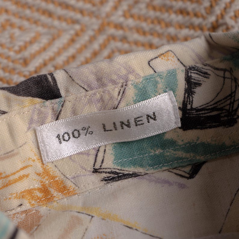 May include: Close-up of a fabric label that reads "100% LINEN." The fabric is a light cream color with a pattern of black, teal, and purple abstract shapes. The label is white with black text.