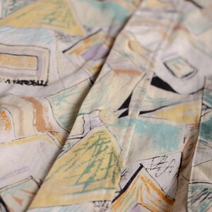 May include: Close-up of a button-down shirt with a colorful abstract pattern. The shirt features a white base with geometric shapes in yellow, blue, purple, and black. The fabric is slightly wrinkled, and a button is visible.
