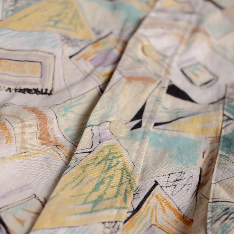 May include: Close-up of a button-down shirt with a colorful abstract pattern. The shirt features a white base with geometric shapes in yellow, blue, purple, and black. The fabric is slightly wrinkled, and a button is visible.