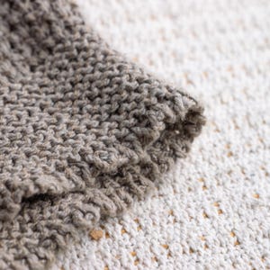 May include: Close-up of a section of knitted fabric in shades of gray. The texture is visible, with the stitches creating a raised pattern. The edge of the fabric is rolled over, revealing the thickness and construction of the knit.