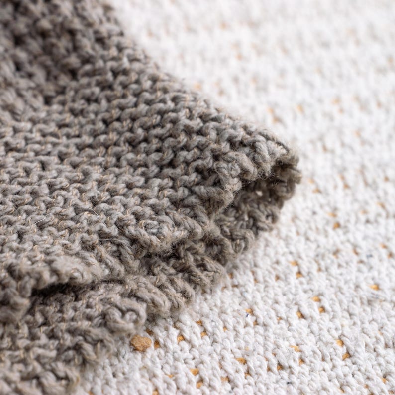 May include: Close-up of a section of knitted fabric in shades of gray. The texture is visible, with the stitches creating a raised pattern. The edge of the fabric is rolled over, revealing the thickness and construction of the knit.