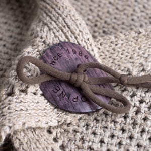 May include: Close-up of a beige knit fabric with a large, round, dark wood button. The button has the text "PINE 1986" engraved on it and is attached with a brown cord loop. The fabric has a textured, open weave.