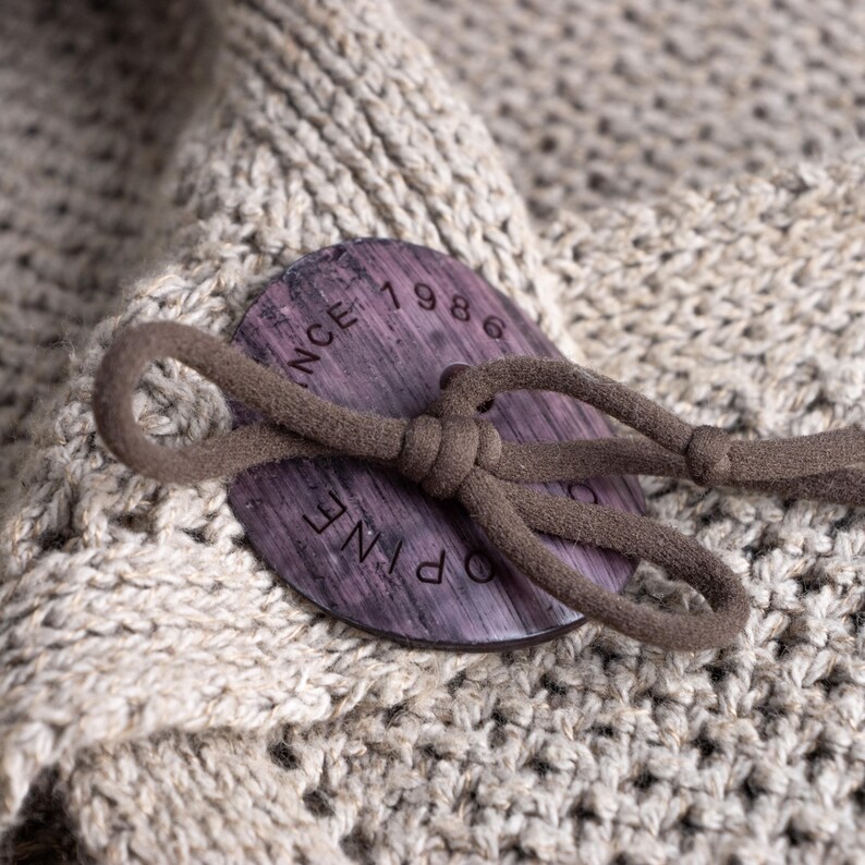May include: Close-up of a beige knit fabric with a large, round, dark wood button. The button has the text "PINE 1986" engraved on it and is attached with a brown cord loop. The fabric has a textured, open weave.