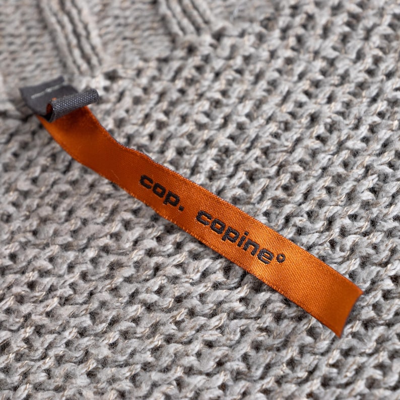 May include: Close-up of a gray knit fabric with a textured pattern. An orange tag with the text "cop. copine" is attached to the fabric. The tag is made of a smooth, satin-like material.