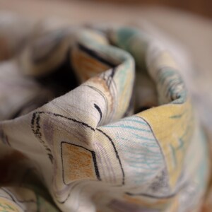 May include: Close-up of a folded fabric with a colorful, abstract pattern. The design features geometric shapes and lines in shades of yellow, blue, orange, and purple on a light background. The fabric appears to be soft and lightweight.