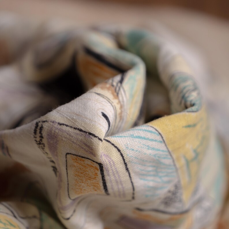May include: Close-up of a folded fabric with a colorful, abstract pattern. The design features geometric shapes and lines in shades of yellow, blue, orange, and purple on a light background. The fabric appears to be soft and lightweight.