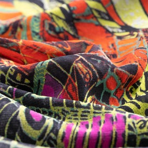 May include: Close-up of a patterned fabric in shades of orange, black, yellow, green, and pink. The design is abstract, with a mix of geometric shapes and artistic elements. The fabric is likely used for clothing or accessories.
