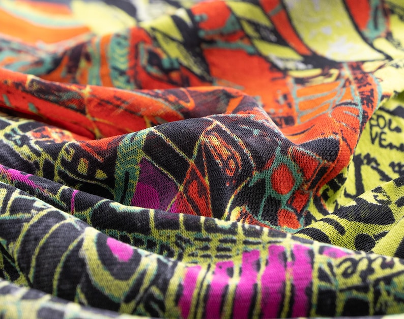 May include: Close-up of a patterned fabric in shades of orange, black, yellow, green, and pink. The design is abstract, with a mix of geometric shapes and artistic elements. The fabric is likely used for clothing or accessories.