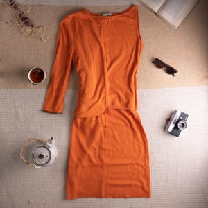 Könnte beinhalten: Ein zweiteiliges Outfit in Orange, bestehend aus einem langärmeligen Oberteil und einem Rock, wird auf einer neutralen Oberfläche präsentiert. Eine kleine Teekanne, eine Tasse Tee, eine Sonnenbrille, eine Vintage-Kamera und ein offenes Buch sind um das Outfit herum angeordnet.