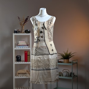 Könnte beinhalten: Ein ärmelloses Patchwork-Kleid auf einer Schaufensterpuppe. Das Kleid weist eine Mischung aus Texturen und Mustern auf, darunter gewebte, gemusterte und unifarbene Stoffe in Beige, Creme und Schwarz. Es hat große Knöpfe und einen Fransenbesatz. Eine silberne Halskette befindet sich auf der Schaufensterpuppe.