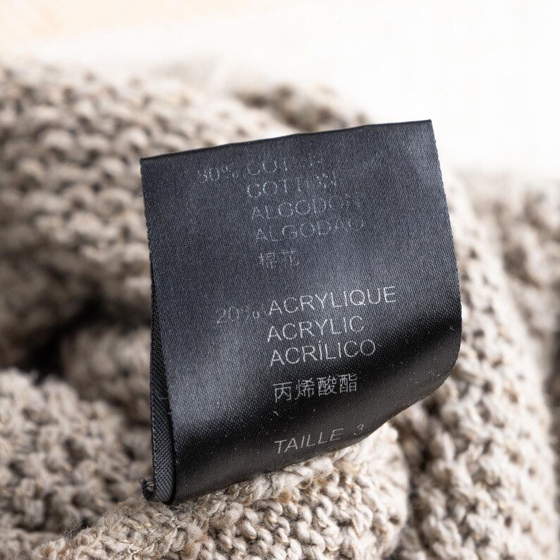 May include: Close-up of a black clothing tag with white text. The tag lists the material composition as 30% cotton and 20% acrylic, with additional text in multiple languages. The tag is attached to a beige knit fabric.