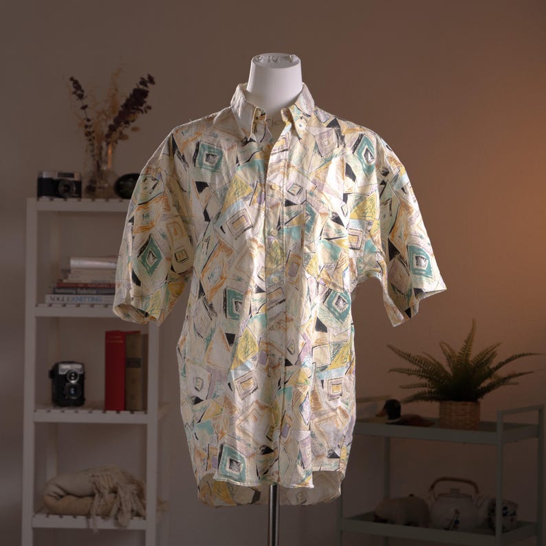 May include: A short-sleeved button-down shirt with a geometric pattern in shades of yellow, green, and black. The shirt is displayed on a mannequin, showcasing its loose fit and retro design. The shirt's collar and cuffs are white.