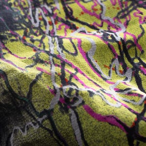 May include: Close-up of a fabric with an abstract design. The pattern has a yellow-green base with black, white, and pink lines and splatters. The fabric appears soft and textured, with visible folds and creases.