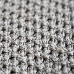 May include: Close-up of a textured, light gray knitted fabric. The fabric has a dense, raised pattern, creating a tactile surface. The yarn appears to be a blend of light and darker gray fibers, giving a heathered effect. The overall look is soft and inviting.