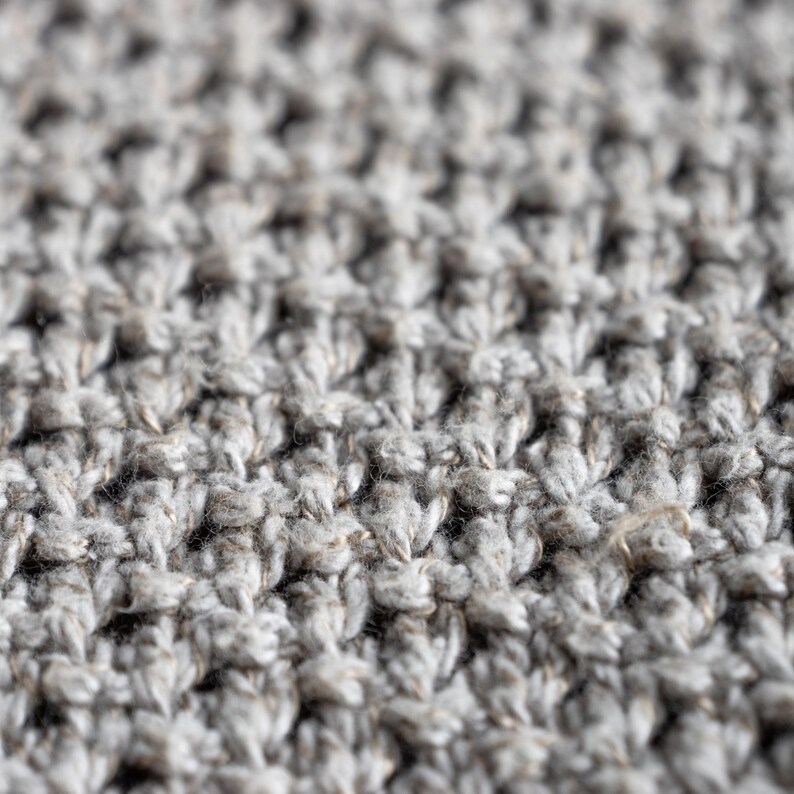 May include: Close-up of a textured, light gray knitted fabric. The fabric has a dense, raised pattern, creating a tactile surface. The yarn appears to be a blend of light and darker gray fibers, giving a heathered effect. The overall look is soft and inviting.