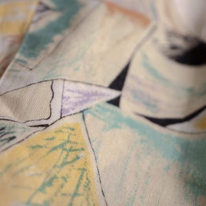 May include: Close-up of a fabric with a pastel abstract design. The pattern features shapes in yellow, light blue, purple, and black, outlined with black lines, set against a cream background. The fabric appears to be soft and textured.