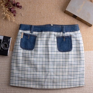 Könnte beinhalten: Minirock aus Jeansstoff und kariertem Stoff mit zwei Vordertaschen. Der Rock hat einen Jeansbund und -taschenakzente sowie ein blau-weißes Karomuster. Eine Vintage-Kamera und ein Buch befinden sich im Hintergrund.