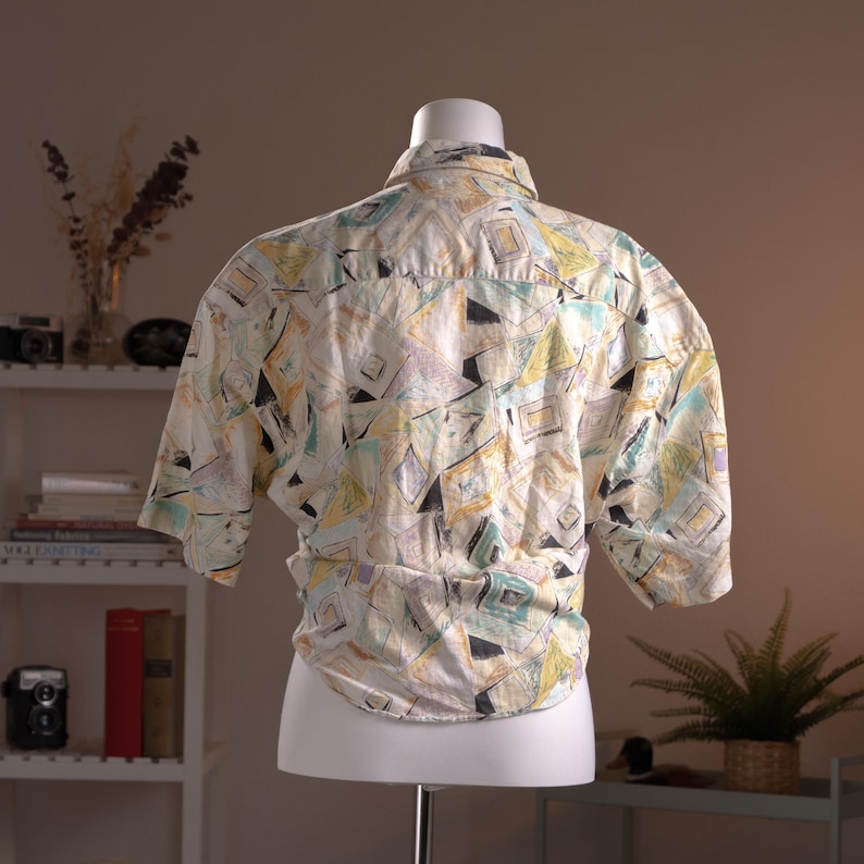 May include: A short-sleeved button-down shirt with a geometric pattern in pastel colors of yellow, blue, and purple on a white background. The shirt is displayed on a mannequin.