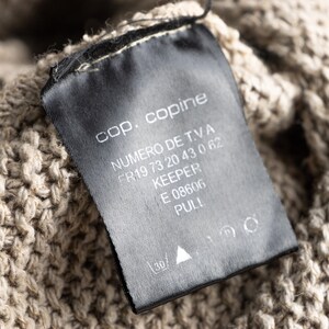 May include: Close-up of a black clothing tag with white text. The tag reads "cop. copine", "NUMERO DE T.V.A", "KEEPER", "E 08606", and "PULL". The tag is attached to a beige knitted fabric.