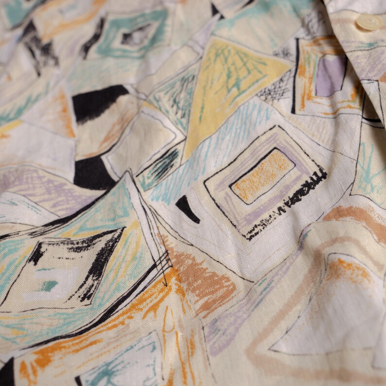 May include: Close-up of a cream-colored fabric with a repeating geometric pattern. The design features squares, rectangles, and triangles in shades of yellow, black, teal, orange, and purple. The fabric appears to be slightly wrinkled.