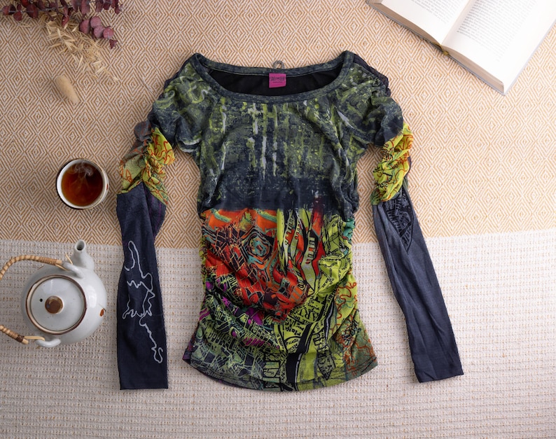 May include: A long-sleeved top with a distinctive, colourful design. The top features a mix of green, orange, and blue hues, with a ruched bodice and sleeves. A cup of tea and a teapot are also visible.