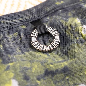 May include: Close-up of a black mesh fabric with a yellow and green abstract pattern. A silver ring with the words "SAVE THE QUEENS" is attached to the fabric with a black ribbon.