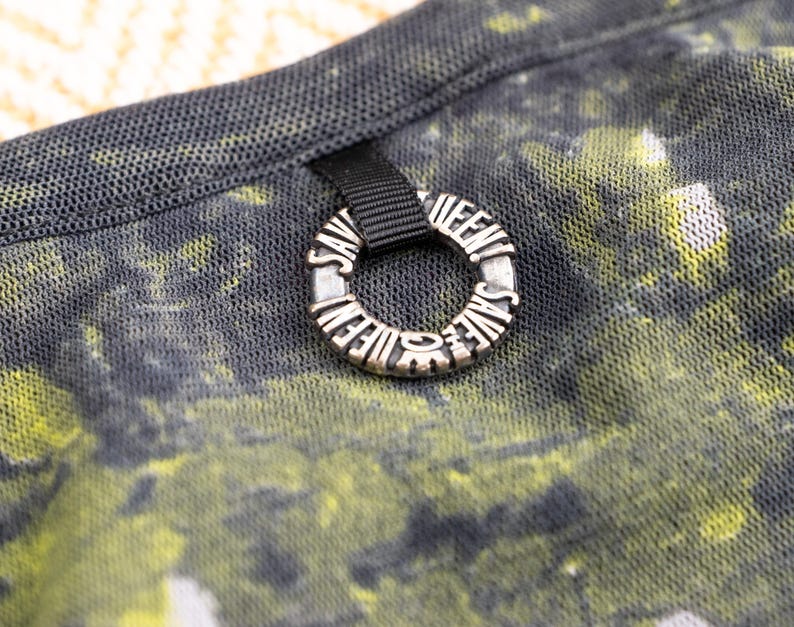 May include: Close-up of a black mesh fabric with a yellow and green abstract pattern. A silver ring with the words "SAVE THE QUEENS" is attached to the fabric with a black ribbon.