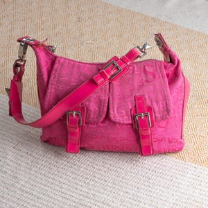 Könnte beinhalten: Eine pinkfarbene Umhängetasche mit einem sich wiederholenden "VERSUS"-Muster. Die Tasche hat einen langen Riemen, zwei Vordertaschen mit silbernen Schnallen und einen silbernen Griffansatz. Die Tasche ist aus weichem Material und vor einem strukturierten Hintergrund platziert.