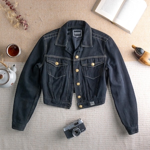 Może przedstawiać: Ciemnoniebieska kurtka dżinsowa ze złotymi guzikami i przeszyciami. Kurtka ma klasyczny kołnierz, dwie kieszenie na piersi i krótki krój. Widoczna jest metka Versace. Na zdjęciu znajduje się również aparat fotograficzny w stylu vintage i filiżanka herbaty.