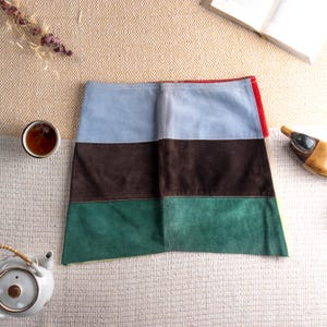 May include: A suede mini skirt featuring horizontal panels in blue, brown, and green. The skirt has a red accent on the side. It is displayed on a textured surface. A unique piece of clothing.