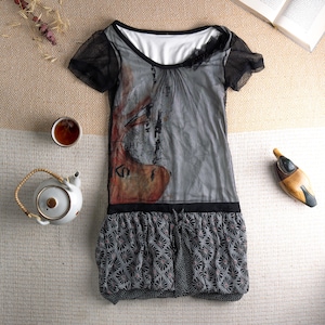 Könnte beinhalten: Ein graues und schwarzes Kleid mit kurzen Ärmeln und gemustertem Rock. Das Kleid hat einen transparenten Überzug mit einem abstrakten Design. Eine weiße Teekanne, eine Tasse Tee und eine Holzvogelfigur sind ebenfalls sichtbar.