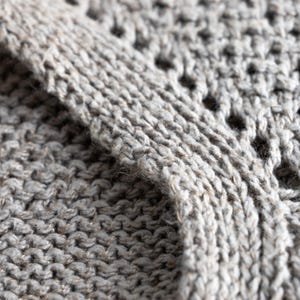 May include: Close-up of a knitted fabric in shades of gray. The texture reveals a combination of solid knit and openwork patterns, creating a visually interesting surface. The yarn appears to be a blend of fibers, adding depth and dimension to the textile.