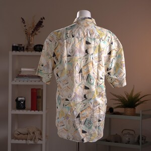 May include: Short-sleeved button-down shirt with a geometric pattern in pastel colors. The design features squares, triangles, and diamond shapes in shades of yellow, blue, purple, and black. The shirt is displayed on a mannequin.