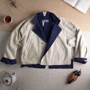 May include: Cream-colored jacket with navy blue accents. The jacket features a contrasting navy blue collar, cuffs, and interior lining. The label inside the jacket reads "WIN". The jacket is buttoned and has two front pockets.