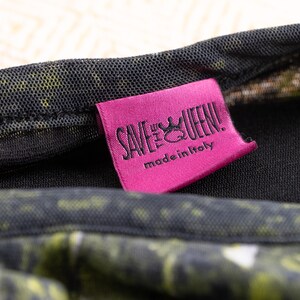 May include: A close-up shot of a pink label with the text "SAVE THE QUEEN!" and "made in Italy" in black. The label is attached to a garment with a black mesh overlay and a green and yellow pattern.