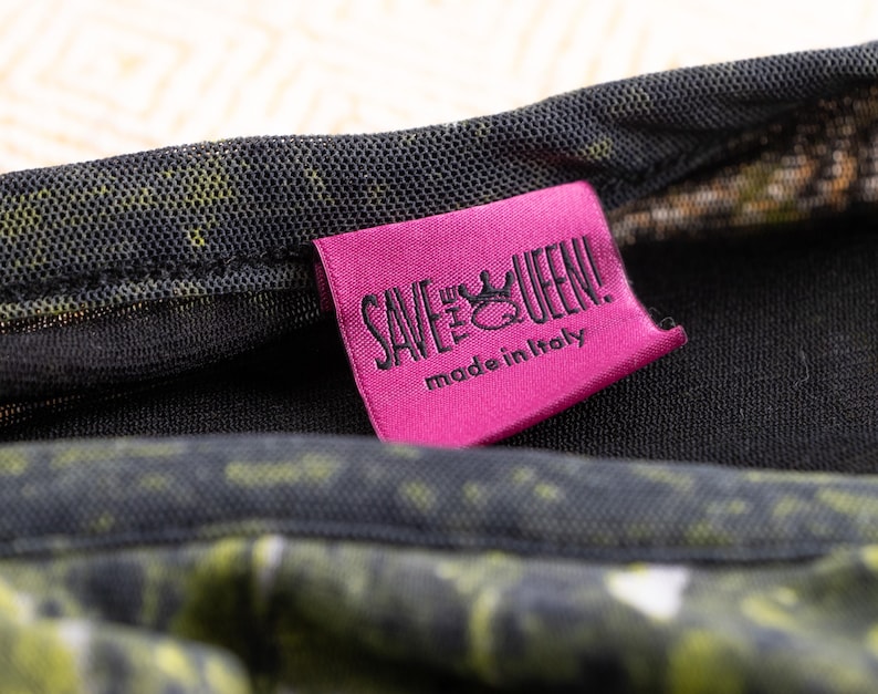 May include: A close-up shot of a pink label with the text "SAVE THE QUEEN!" and "made in Italy" in black. The label is attached to a garment with a black mesh overlay and a green and yellow pattern.